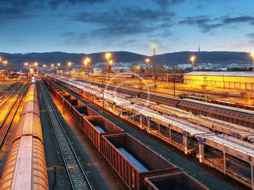 Railway yard at dusk with illuminated tracks, cargo trains, and industrial buildings in the background.