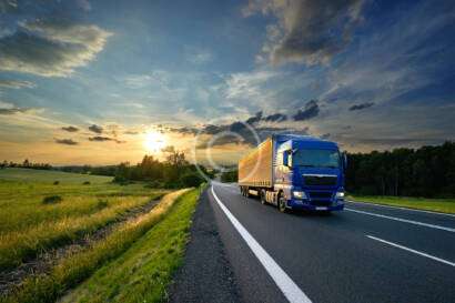 A blue truck drives on a rural highway at sunset, with fields and trees on either side under a partly cloudy sky.