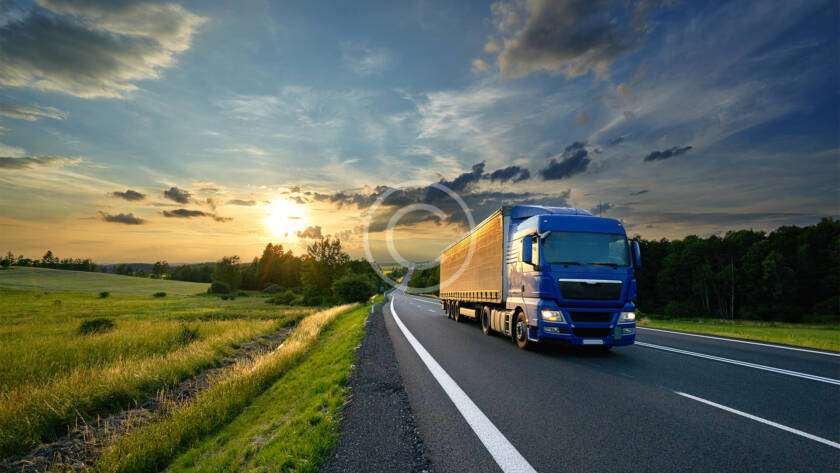 A blue truck drives on a rural highway at sunset, with fields and trees on either side under a partly cloudy sky.