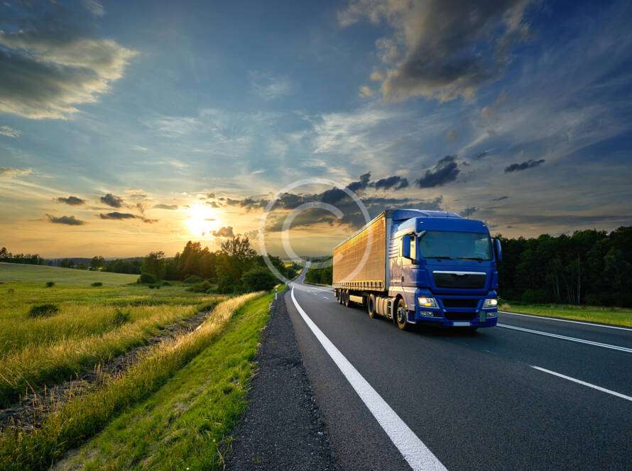 A blue truck drives on a rural highway at sunset, with fields and trees on either side under a partly cloudy sky.