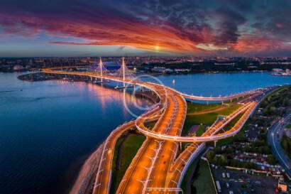 Aerial view of a brightly lit suspension bridge over a river at sunset, with a cityscape in the background and dramatic clouds in the sky.