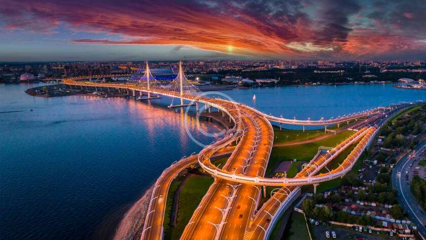 Aerial view of a brightly lit suspension bridge over a river at sunset, with a cityscape in the background and dramatic clouds in the sky.