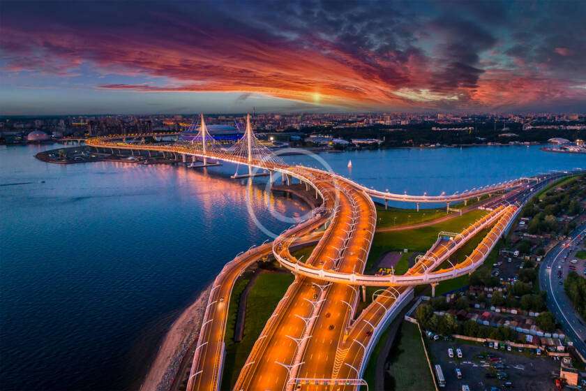 Aerial view of a brightly lit suspension bridge over a river at sunset, with a cityscape in the background and dramatic clouds in the sky.