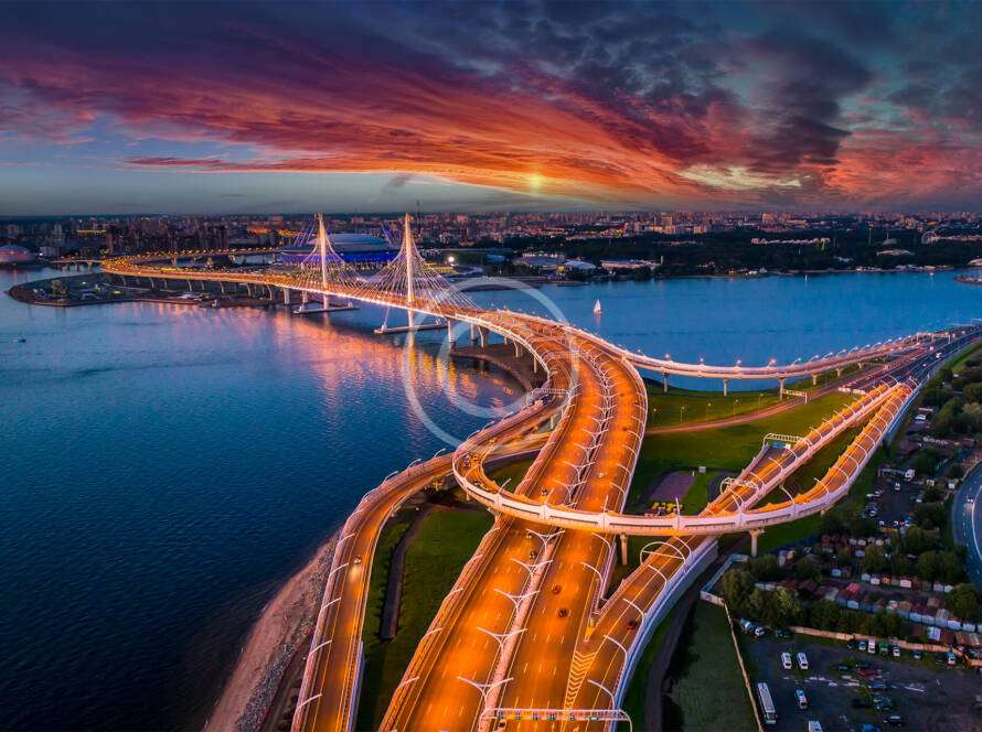 Aerial view of a brightly lit suspension bridge over a river at sunset, with a cityscape in the background and dramatic clouds in the sky.