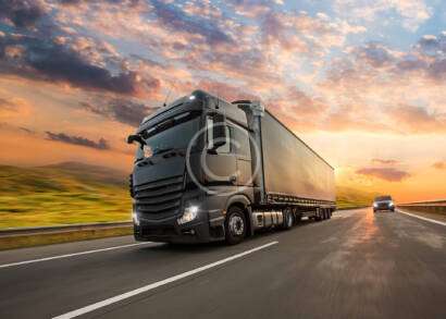 Two trucks drive on a highway during a colorful sunset, with grassy fields lining the road.
