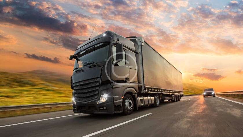 Two trucks drive on a highway during a colorful sunset, with grassy fields lining the road.