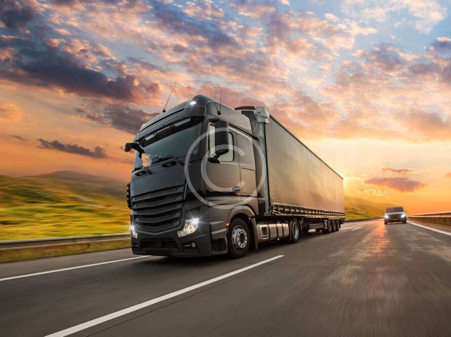 Two trucks drive on a highway during a colorful sunset, with grassy fields lining the road.