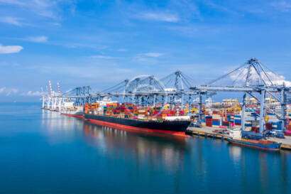 A cargo ship is docked at a busy port, surrounded by cranes and colorful shipping containers under a clear blue sky.