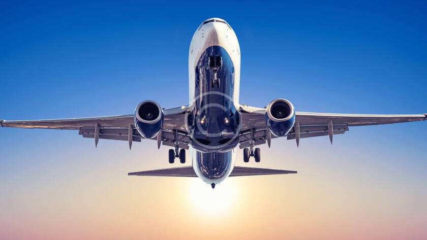 Airplane flying directly overhead against a clear blue sky with the sun setting in the background.