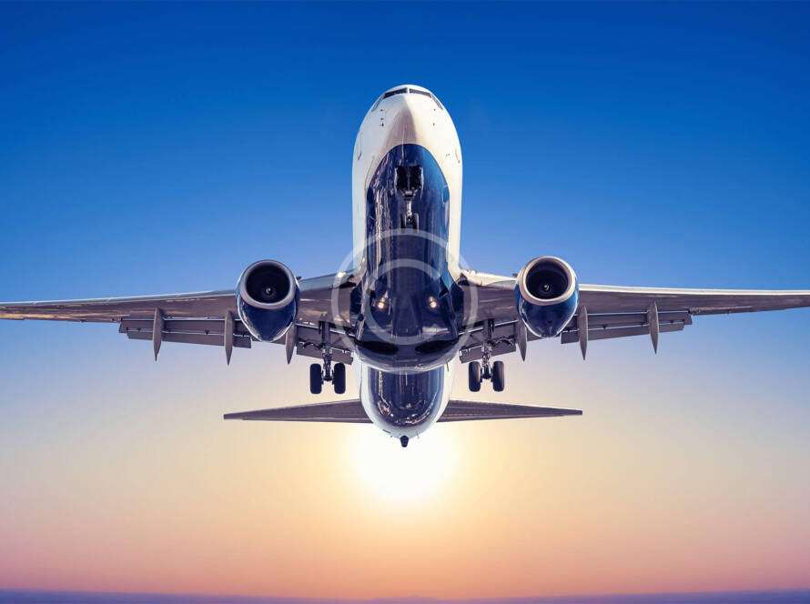 Airplane flying directly overhead against a clear blue sky with the sun setting in the background.