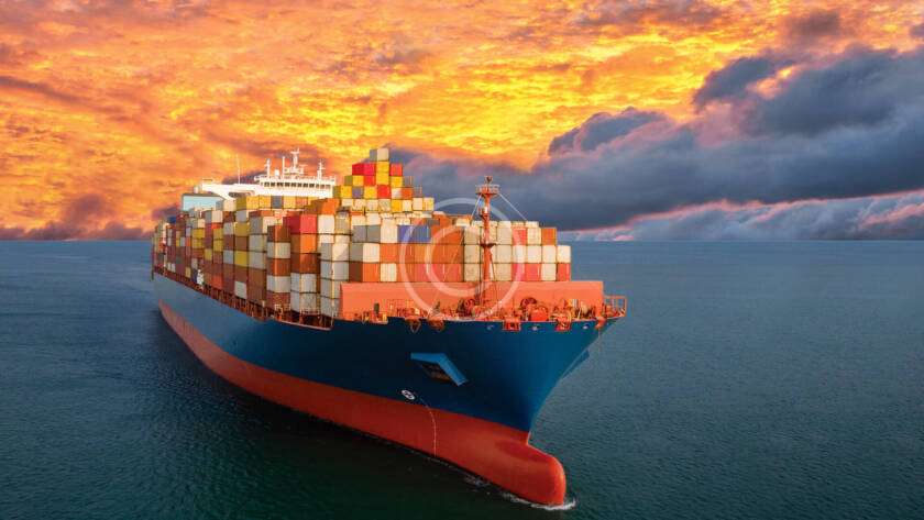 A large cargo ship loaded with colorful containers sails on calm ocean waters under a dramatic orange sunset sky.