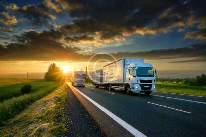 Two trucks drive on a highway at sunset, surrounded by open fields and a partly cloudy sky.