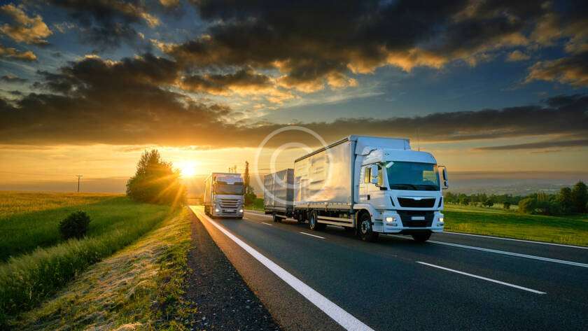 Two trucks drive on a highway at sunset, surrounded by open fields and a partly cloudy sky.