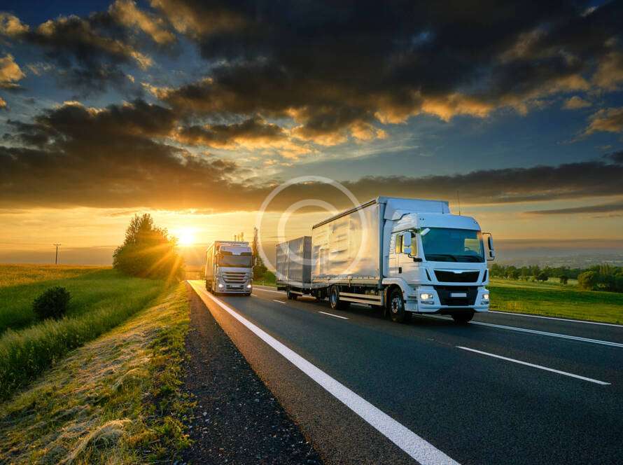 Two trucks drive on a highway at sunset, surrounded by open fields and a partly cloudy sky.