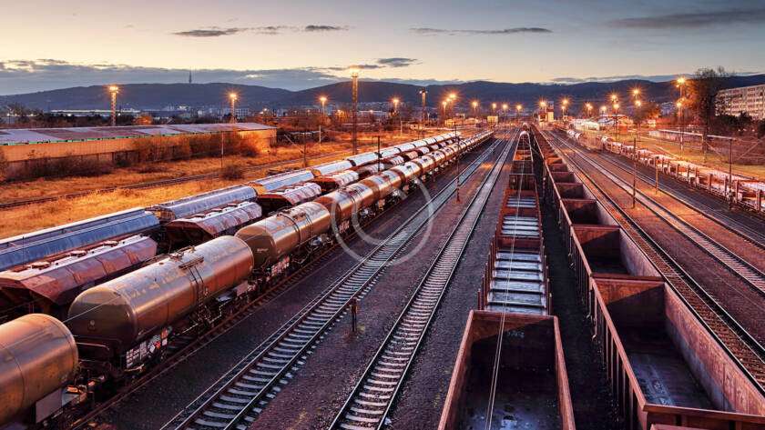 A train yard at dusk with multiple tracks and rows of freight trains. The sky is clear with a few clouds, and industrial buildings are visible in the background.