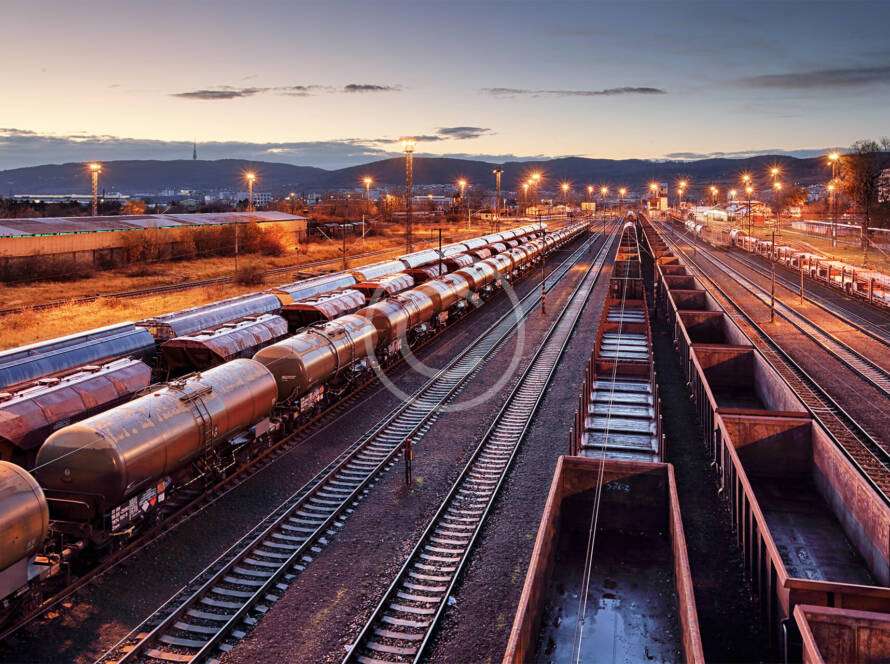 A train yard at dusk with multiple tracks and rows of freight trains. The sky is clear with a few clouds, and industrial buildings are visible in the background.