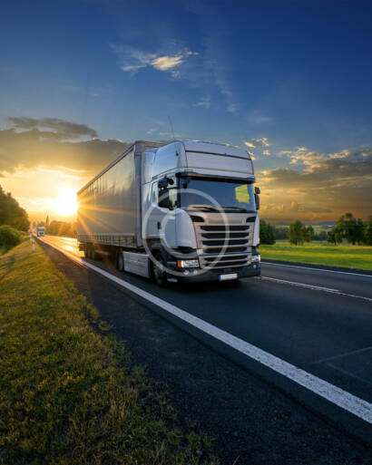 A large truck drives down a rural highway at sunset, with green fields and a clear sky in the background.