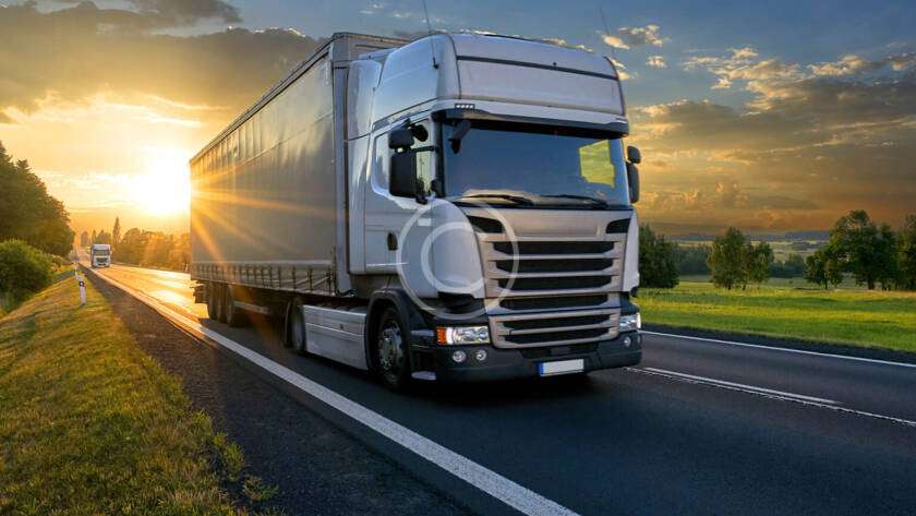 A large truck drives down a rural highway at sunset, with green fields and a clear sky in the background.