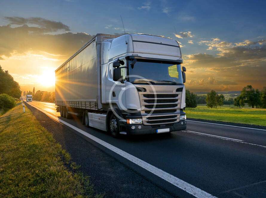 A large truck drives down a rural highway at sunset, with green fields and a clear sky in the background.