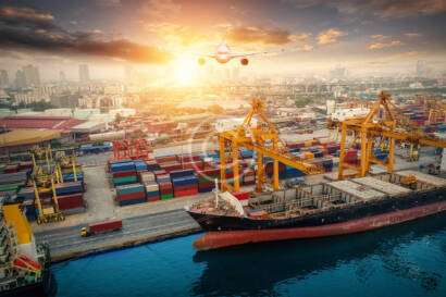 A cargo ship docked at a busy port with colorful shipping containers. A plane flies overhead with a city skyline in the background during sunset.