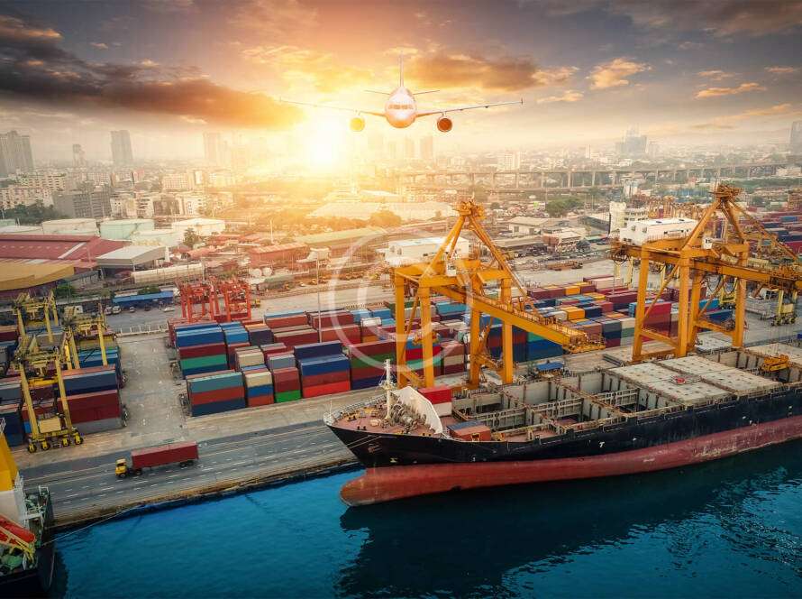A cargo ship docked at a busy port with colorful shipping containers. A plane flies overhead with a city skyline in the background during sunset.