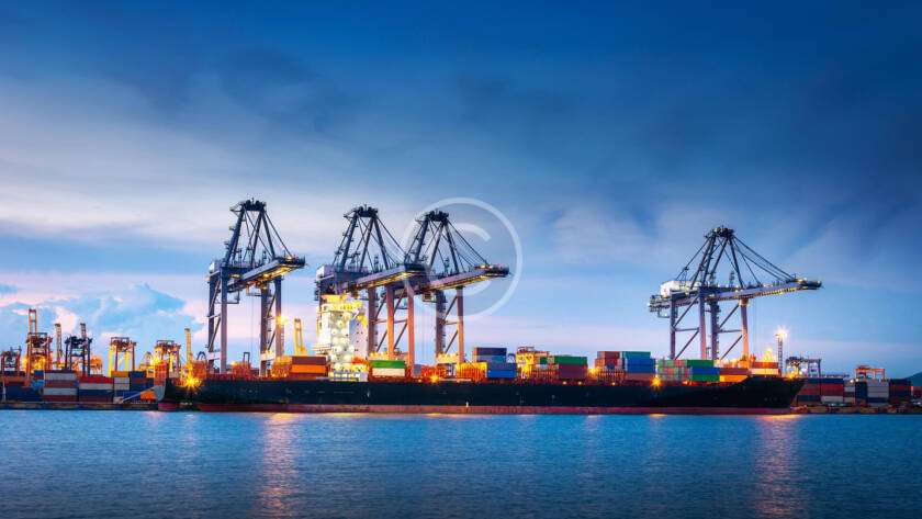 Container ship docked at an illuminated port with cranes, under a dusk sky, surrounded by water.