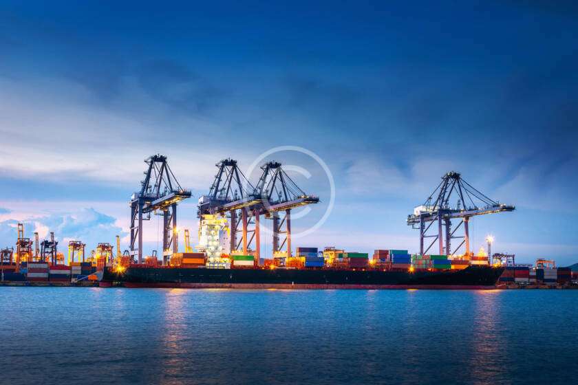Container ship docked at an illuminated port with cranes, under a dusk sky, surrounded by water.
