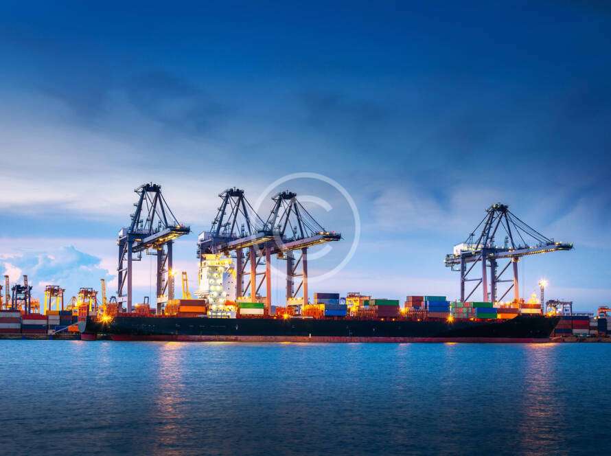 Container ship docked at an illuminated port with cranes, under a dusk sky, surrounded by water.