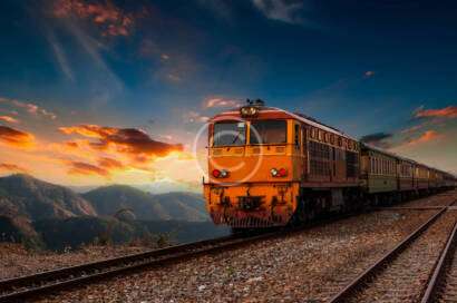 A train travels on tracks through a mountainous landscape at sunset, with vibrant clouds in the sky.
