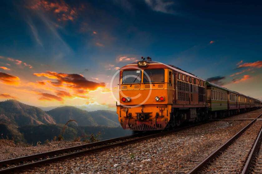 A train travels on tracks through a mountainous landscape at sunset, with vibrant clouds in the sky.