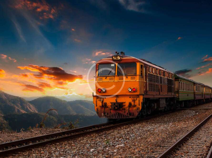 A train travels on tracks through a mountainous landscape at sunset, with vibrant clouds in the sky.