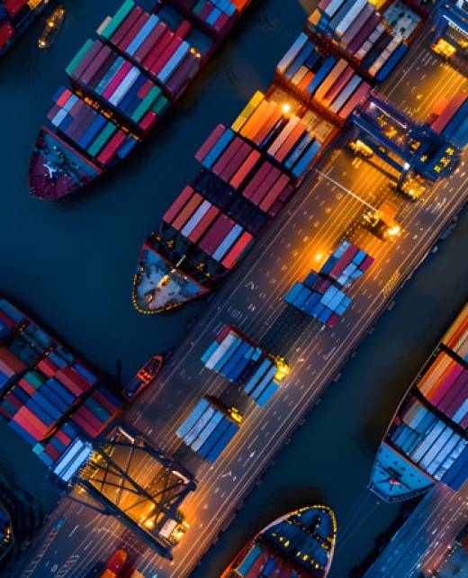 Aerial view of a busy port with cargo ships and containers on both sides of a waterway, illuminated by streetlights at night.
